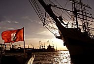 011_15708 Sonnenuntergang im Hamburger Hafen; die Hamburg Fahne strahlt in der Abend Sonne - vom Grosssegler am Kai der Landungsbrücken sind Bug und Masten in der Silhouette zu erkennen - im Hintergrund Kräne der Werft an der Elbe.