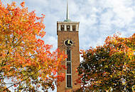 9469 Kirchturm der Frohbotschaftskirche in Hamburg Dulsberg zwischen Herbstbäumen - Blätter herbstlich rot, braun und gelb gefärbt.