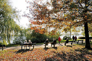 2010 Die Herbstsonne scheint durch das Laub der Bäume am Alsterufer von Hamburg Harvestehude - Spaziergänger gehen auf dem Alsterweg - andere Besucher der Grünanlage sitzen auf den Ruhebänken am Ufer der Aussenalster.