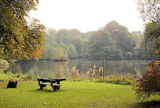 5734 Herbstlich gefärbte Bäume am Mühlenteich in Grosshansdorf; Herbstfarben. Sitzbank / Sitzgruppe aus Holz, Picknickplatz auf einer Wiese am Wasser.