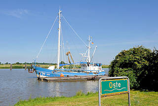 5106 Schild Oste - grüner Grund mit weisser Schrift an der Oste bei Neuhaus - ein blauer Fischkutter liegt am Ufer.